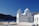 A white building with blue accents and a dome is situated near a coastline, with rocky hills in the background under a clear blue sky. The building features an arched door, several windows, and a striped flagpole with the Greek flag.