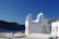 A white building with blue accents and a dome is situated near a coastline, with rocky hills in the background under a clear blue sky. The building features an arched door, several windows, and a striped flagpole with the Greek flag.