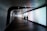A futuristic, curved hallway with illuminated walls displays a gradient of colors. Two people walk together in silhouette along the dimly lit corridor, creating a sense of mystery and intrigue. The ceiling features geometric patterns with recessed lighting.