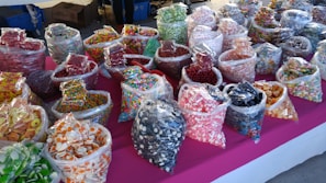 An assortment of various colorful candies displayed in clear plastic bags on a bright pink tablecloth. The bags contain different types of sweets such as gummies, hard candies, and chocolate-covered treats, organized in rows and visually appealing due to the vivid colors and variety.