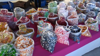 An assortment of various colorful candies displayed in clear plastic bags on a bright pink tablecloth. The bags contain different types of sweets such as gummies, hard candies, and chocolate-covered treats, organized in rows and visually appealing due to the vivid colors and variety.