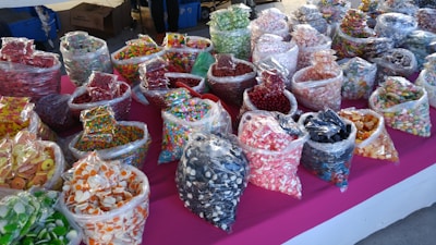 An assortment of various colorful candies displayed in clear plastic bags on a bright pink tablecloth. The bags contain different types of sweets such as gummies, hard candies, and chocolate-covered treats, organized in rows and visually appealing due to the vivid colors and variety.
