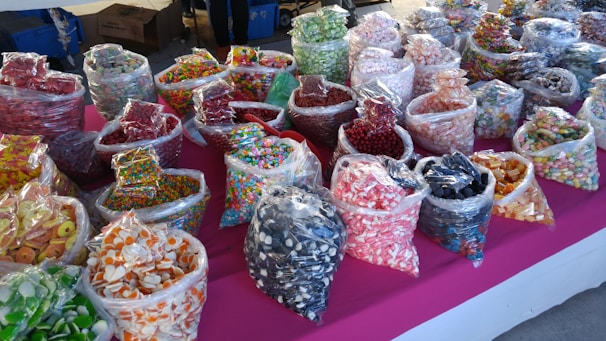 An assortment of various colorful candies displayed in clear plastic bags on a bright pink tablecloth. The bags contain different types of sweets such as gummies, hard candies, and chocolate-covered treats, organized in rows and visually appealing due to the vivid colors and variety.