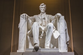 A large marble statue of a seated man, depicting an iconic historical figure, positioned in a grand architectural setting with columns and shadows.