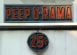 A vintage-style sign reading 'PEEP O RAMA' in large, bold red letters against a black background. Below it, a circular sign displays '25¢' in a similar style. Both signs have a retro, illuminated appearance.