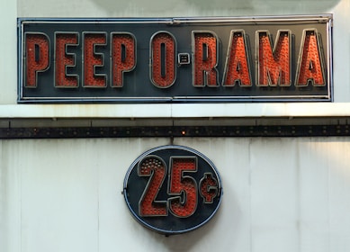 A vintage-style sign reading 'PEEP O RAMA' in large, bold red letters against a black background. Below it, a circular sign displays '25¢' in a similar style. Both signs have a retro, illuminated appearance.