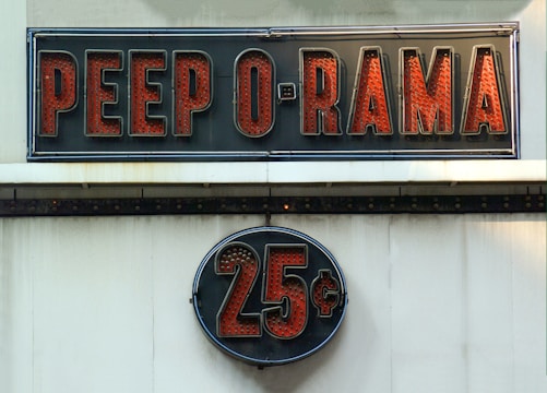 A vintage-style sign reading 'PEEP O RAMA' in large, bold red letters against a black background. Below it, a circular sign displays '25¢' in a similar style. Both signs have a retro, illuminated appearance.