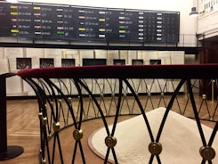 A room with a dark display board filled with handwritten chalk listings dominates the background. Below it, large open books and historical photographs are on display. In the foreground, a metallic barrier with a red velvet top surrounds a mound of light-colored material.