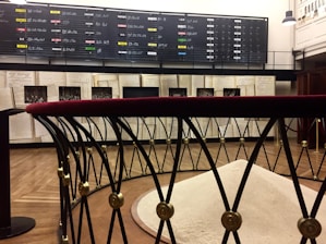 A room with a dark display board filled with handwritten chalk listings dominates the background. Below it, large open books and historical photographs are on display. In the foreground, a metallic barrier with a red velvet top surrounds a mound of light-colored material.