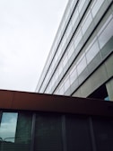A modern architectural structure with a series of large glass windows aligned in a grid, creating a reflective and sleek appearance. The building has a combination of metal and glass surfaces, with the lower section featuring darker panels.