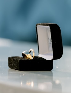 A gold ring is elegantly placed inside an open black velvet ring box. The box sits on a reflective surface, and the background is softly blurred in a calming blue color.
