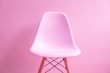 A modern, smooth plastic chair with wooden legs, set against a monochromatic pink background. The chair’s sleek design contrasts subtly with the textured wall.