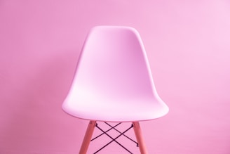 A modern, smooth plastic chair with wooden legs, set against a monochromatic pink background. The chair’s sleek design contrasts subtly with the textured wall.