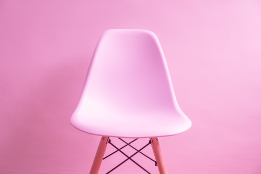 A modern, smooth plastic chair with wooden legs, set against a monochromatic pink background. The chair’s sleek design contrasts subtly with the textured wall.