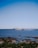 A ferry labeled 'coloniaexpress' moves across a calm body of water under a clear blue sky. In the foreground, there is a rocky shore with patches of green vegetation.