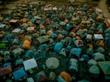 A densely packed array of colorful tents spans across a large outdoor area, suggesting a crowded campsite setting with various makeshift shelters. In the background, a few buses and vehicles are parked, indicating temporary accommodations and transport options.