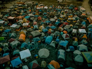 A densely packed array of colorful tents spans across a large outdoor area, suggesting a crowded campsite setting with various makeshift shelters. In the background, a few buses and vehicles are parked, indicating temporary accommodations and transport options.