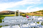 A serene outdoor scene set on a grassy hilltop with rows of white chairs arranged for what appears to be a wedding ceremony. An arbor decorated with flowers is centered in the background under a bright blue sky with wispy clouds. The landscape features rolling hills and sparse trees, contributing to the calm and picturesque setting.