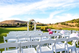 A serene outdoor scene set on a grassy hilltop with rows of white chairs arranged for what appears to be a wedding ceremony. An arbor decorated with flowers is centered in the background under a bright blue sky with wispy clouds. The landscape features rolling hills and sparse trees, contributing to the calm and picturesque setting.