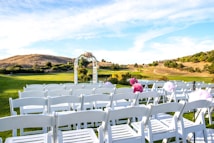 A serene outdoor scene set on a grassy hilltop with rows of white chairs arranged for what appears to be a wedding ceremony. An arbor decorated with flowers is centered in the background under a bright blue sky with wispy clouds. The landscape features rolling hills and sparse trees, contributing to the calm and picturesque setting.