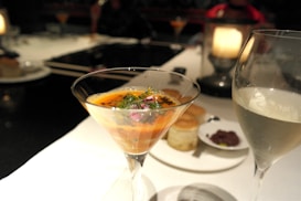 A sophisticated dining setup featuring a cocktail or dessert garnished with herbs in a martini glass. Beside it, a glass of white wine or champagne is seen. The table is elegantly set with white tablecloths, and in the background, there are blurred images of additional diners and ambient lighting, creating a warm and welcoming atmosphere.