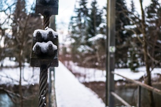 A close-up of a metal cable with a clamp covered in snow. The background features a snow-covered bridge leading into a forested area with leafless trees and evergreens.