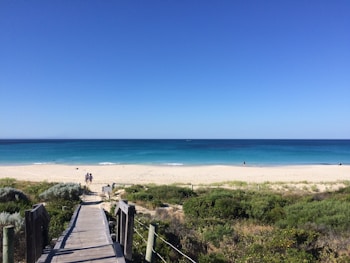 A wooden boardwalk leads to a sandy beach with vibrant blue ocean waters stretching to the horizon. Sparse vegetation surrounds the boardwalk, and a few people are visible walking and enjoying the beach.