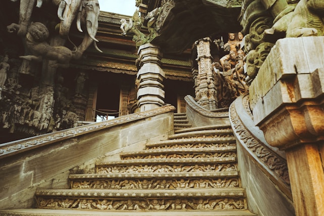 Intricately carved wooden staircase leads to a temple structure adorned with detailed sculptures and ornate designs, featuring mythical and religious figures. The architecture displays elaborate craftsmanship with various animals and deities embedded in the carvings.