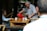 An elderly person is seated at a wooden table outdoors, while another elderly person pours liquid from a red thermos into a cup. The setting appears to be a social one with multiple people in the background, partially obscured by trees and other structures.