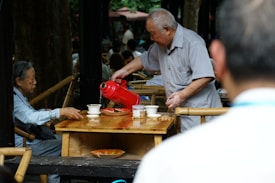 An elderly person is seated at a wooden table outdoors, while another elderly person pours liquid from a red thermos into a cup. The setting appears to be a social one with multiple people in the background, partially obscured by trees and other structures.