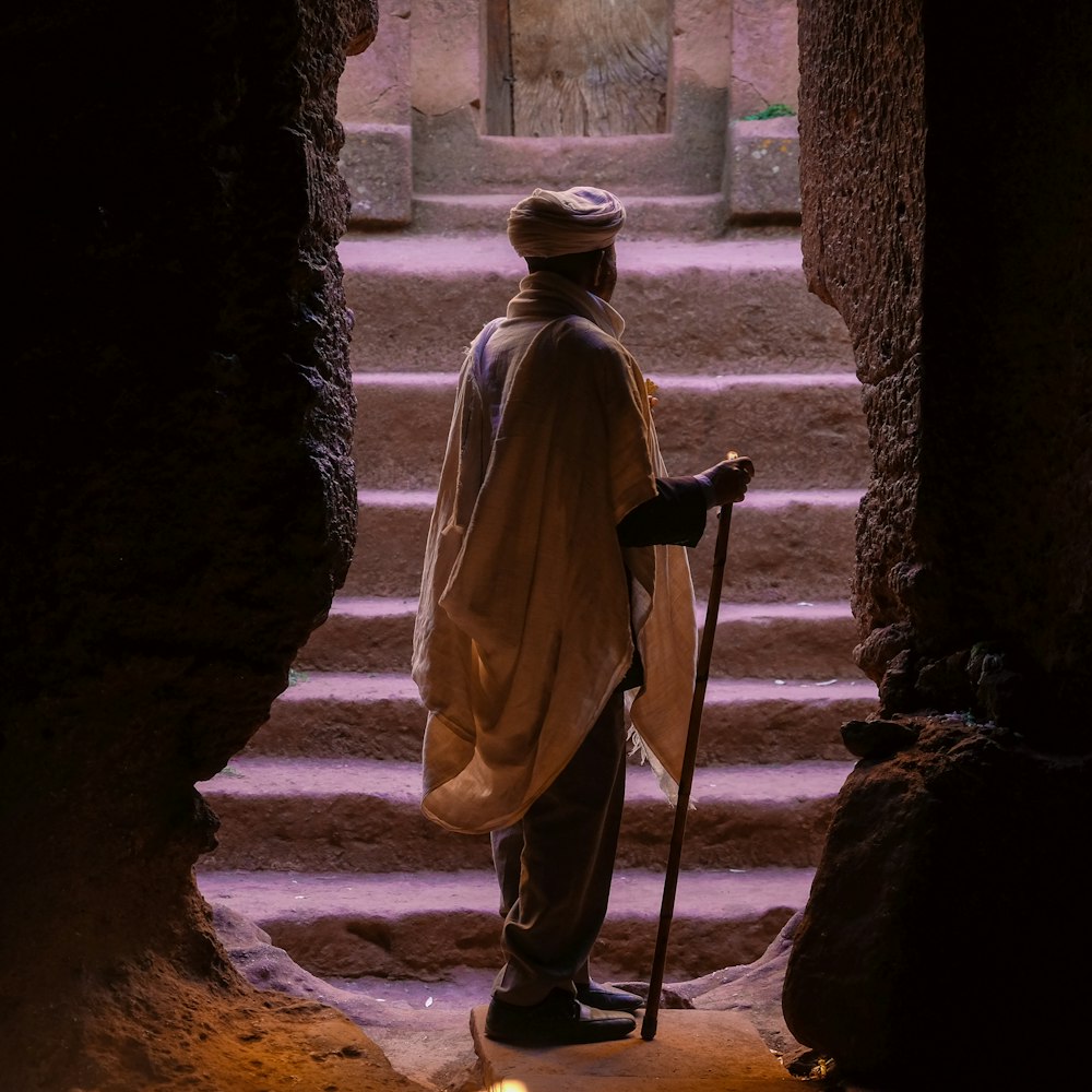 지팡이를 들고 갈색 돌 동굴 입구에 서 있는 남자