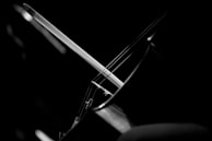 A black and white close-up of a cello featuring the strings and part of the bow. The composition focuses on the interaction between the bow and strings, highlighting the curves and lines of the instrument against a dark background.
