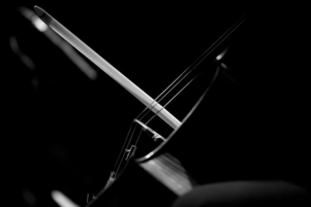 A black and white close-up of a cello featuring the strings and part of the bow. The composition focuses on the interaction between the bow and strings, highlighting the curves and lines of the instrument against a dark background.