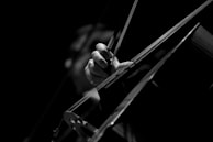 A black and white close-up of a person's hand playing a cello. The hand is gripping the bow, drawing it across the strings. The lighting is focused and dramatic, highlighting the hand and instrument while the background remains dark.