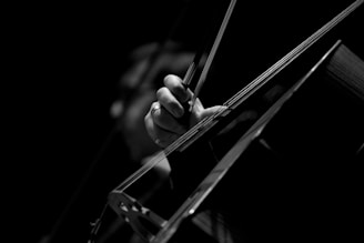 A black and white close-up of a person's hand playing a cello. The hand is gripping the bow, drawing it across the strings. The lighting is focused and dramatic, highlighting the hand and instrument while the background remains dark.