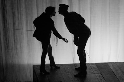 The lead actors sharing an intense moment on stage with charcoal-toned backdrops and textured set design.