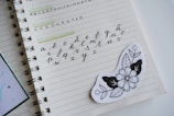 A notebook is open to a page displaying various types of lettering exercises. The page contains uppercase and lowercase alphabets in different writing styles, including brush lettering. An artistic black and white floral sticker is placed on the page.
