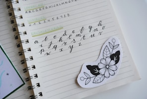 A notebook is open to a page displaying various types of lettering exercises. The page contains uppercase and lowercase alphabets in different writing styles, including brush lettering. An artistic black and white floral sticker is placed on the page.