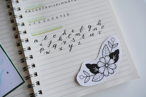A notebook is open to a page displaying various types of lettering exercises. The page contains uppercase and lowercase alphabets in different writing styles, including brush lettering. An artistic black and white floral sticker is placed on the page.