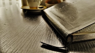 A leather-bound notebook is placed on a glossy wooden table with a ribbon bookmark peeking out. In the background, a cup on a saucer is partially visible, possibly containing coffee or tea, and a blurred object, which might be a bowl or another item, can be seen.