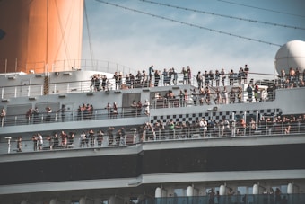 A large cruise ship filled with numerous people standing on several decks, enjoying the view. The ship has a large orange smokestack and a prominent checkered design along one of its sections. The sky in the background is blue and slightly cloudy.