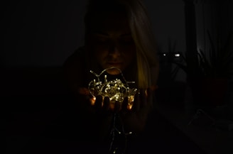 A person holds a cluster of glowing string lights close to their face in a dimly lit room, creating a warm illumination against the darkness. The ambient light highlights parts of the person's face and hands while keeping the surroundings in shadow. Potted plants are vaguely visible in the background, adding a subtle natural element.