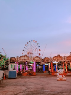 A vibrant amusement park scene featuring a large Ferris wheel with multicolored cabins in the background. The foreground has a carnival booth decorated with ornate patterns and lit with colorful lights. A large bear statue is positioned prominently on the right side. The sky is a soft gradient of blue and pink, creating a serene and lively atmosphere.