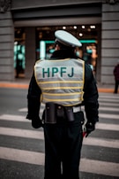A person wearing a reflective vest with 'HPFJ' written on the back, standing on a street with a pedestrian crossing. The individual is dressed in a dark uniform and a white cap, with additional gear around their waist. The background shows a building with bright lights and windows.