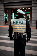 A person wearing a reflective vest with 'HPFJ' written on the back, standing on a street with a pedestrian crossing. The individual is dressed in a dark uniform and a white cap, with additional gear around their waist. The background shows a building with bright lights and windows.