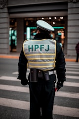 A person wearing a reflective vest with 'HPFJ' written on the back, standing on a street with a pedestrian crossing. The individual is dressed in a dark uniform and a white cap, with additional gear around their waist. The background shows a building with bright lights and windows.