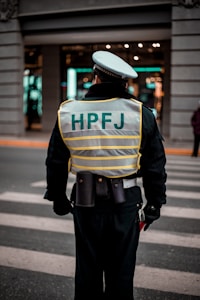 A person wearing a reflective vest with 'HPFJ' written on the back, standing on a street with a pedestrian crossing. The individual is dressed in a dark uniform and a white cap, with additional gear around their waist. The background shows a building with bright lights and windows.