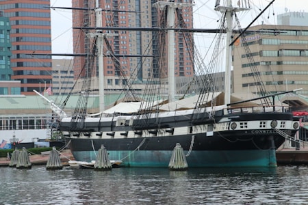 A historic sailing ship is docked at a waterfront area, surrounded by modern high-rise buildings. The ship has multiple masts with numerous ropes and rigging, and the hull is painted in black and white. Several small flags are visible on the ship, and the water in the foreground reflects the structures.