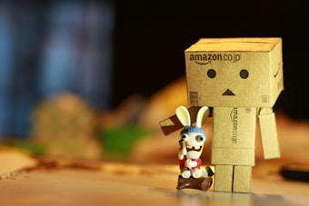 A cardboard robot figurine with a simple, boxy design, featuring a neutral expression made of black circles and a triangle. Next to it stands a small toy rabbit with exaggerated facial features, wearing a red outfit and a blue beanie. The rabbit appears to be holding a wooden club while standing on a small log. The background is blurred, creating a contrast with the sharply focused figures in the foreground.