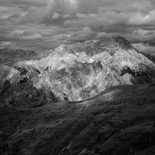 A dramatic black and white photograph of a rugged mountain range under a cloud-filled sky. The peaks are sharply defined with contrasting light and shadow, adding depth and texture to the scene. The clouds above add a sense of impending weather and movement.